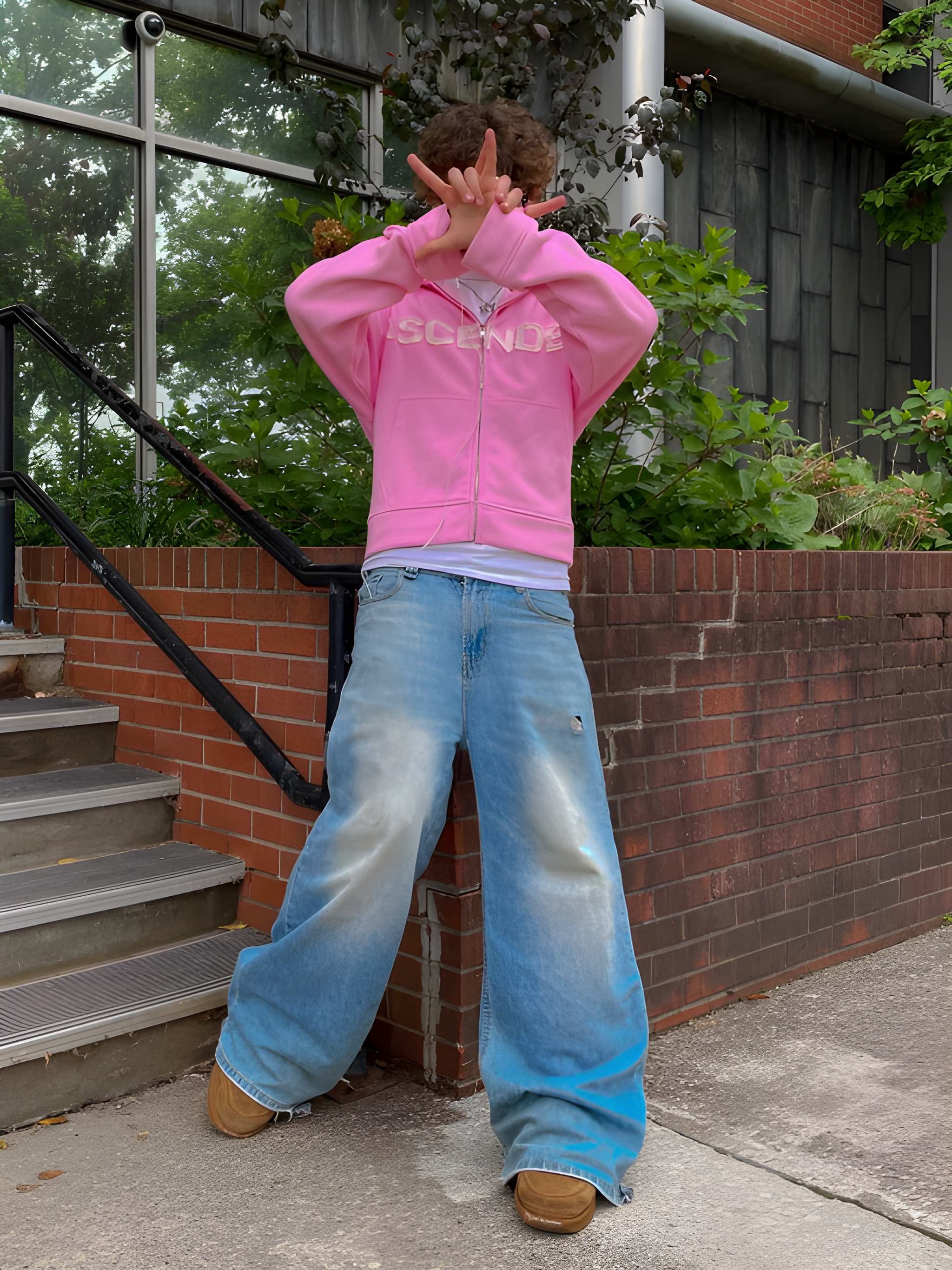 Person wearing a pink hoodie and blue jeans standing on a sidewalk with a building in the background.